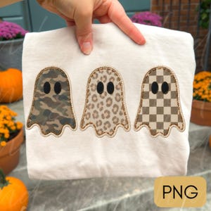 May include: A white t-shirt featuring three embroidered ghost shapes arranged in a row. The first ghost has a green camouflage pattern, the second has a tan leopard print, and the third has a brown and white checkered pattern.