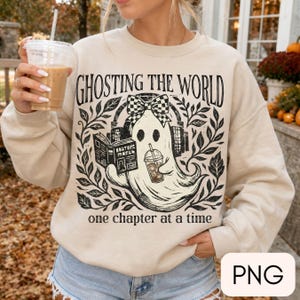 May include: A beige crewneck sweatshirt featuring a black graphic print of a ghost wearing a checkered bow and headphones, reading a book, and holding an iced coffee. The text reads GHOSTING THE WORLD one chapter at a time, surrounded by leaves.