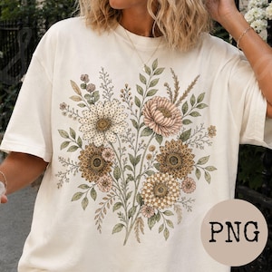 May include: An off-white short-sleeve t-shirt featuring a central graphic of a floral bouquet. The design includes various flowers with intricate patterns, such as polka dots and checkered textures, in shades of brown, beige, and muted green.