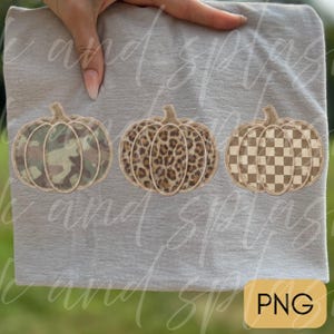 May include: A light gray t-shirt featuring three embroidered pumpkin designs arranged in a horizontal row. The first pumpkin has a green camouflage pattern, the second has a brown leopard print, and the third has a tan and white checkerboard pattern.