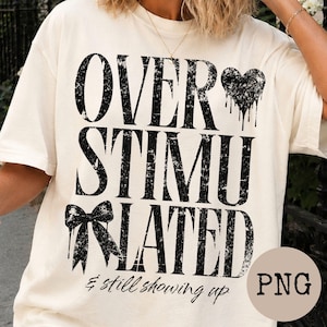 Puede incluir: Una camiseta de color crema con texto negro desgastado que dice OVER STIMULATED, junto a un icono de corazón goteando y un lazo, seguido del texto en cursiva & still showing up. Una insignia circular en la parte inferior derecha muestra el texto PNG.