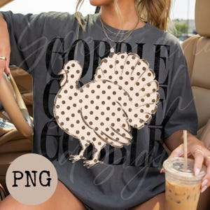 May include: A charcoal gray short-sleeve t-shirt featuring a large graphic of a turkey with a tan body and brown polka dots. The background text reads GOBBLE in a distressed black font. The shirt has a crew neckline and a relaxed fit design.