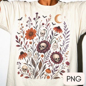Pode incluir: Uma t-shirt de cor creme com uma impressão gráfica detalhada de várias flores silvestres, pequenos crânios no centro das flores, figuras fantasmagóricas, morcegos e uma lua crescente. A ilustração utiliza tons de laranja, roxo e castanho.