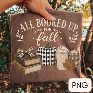 May include: A brown t-shirt featuring the white text ALL BOOKED UP FOR fall, a stack of two books with a plaid bow, a black and white harlequin patterned pumpkin, a cup of coffee with whipped cream, a dried maple leaf, and a plaid candle.