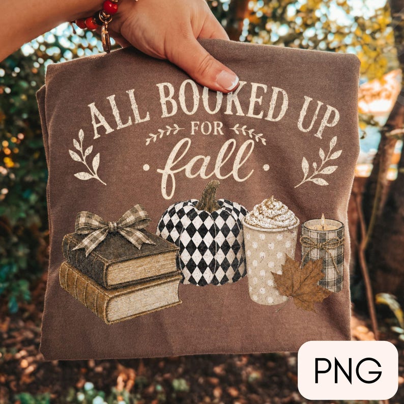 May include: A brown t-shirt featuring the white text ALL BOOKED UP FOR fall, a stack of two books with a plaid bow, a black and white harlequin patterned pumpkin, a cup of coffee with whipped cream, a dried maple leaf, and a plaid candle.