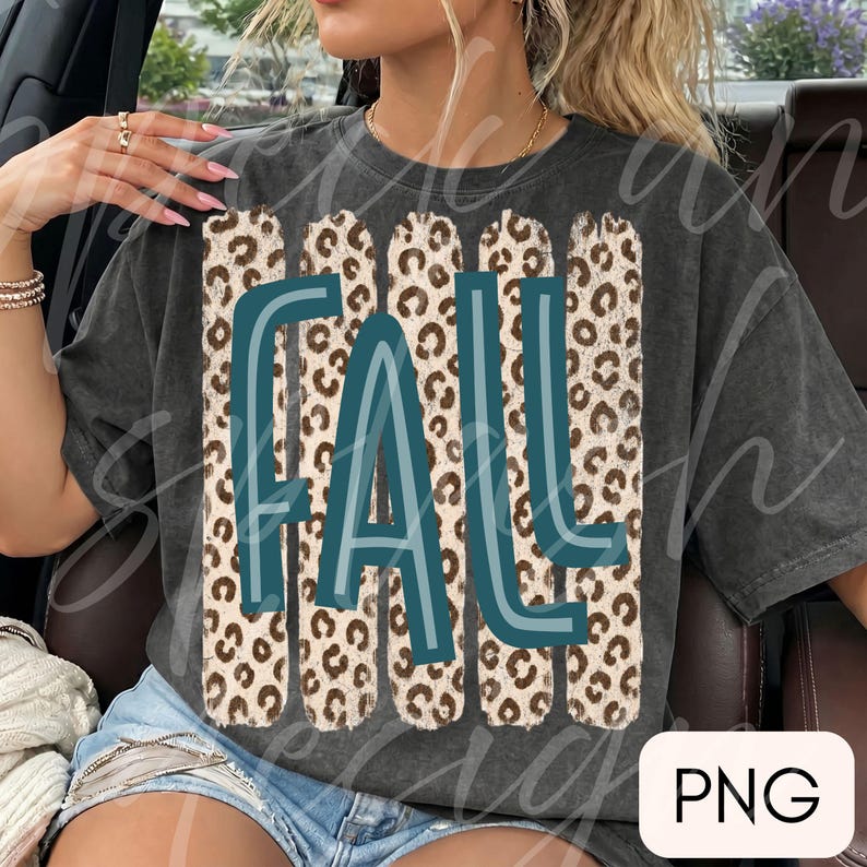May include: A charcoal gray short-sleeve t-shirt featuring a graphic design with the word FALL in teal blue letters. The text is layered over four vertical rectangular stripes with a brown and black leopard print pattern on the fabric.
