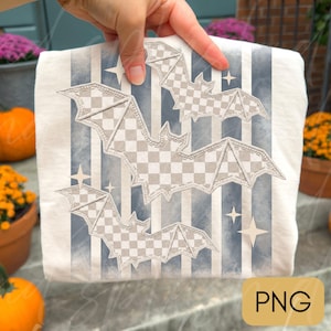 May include: A white t-shirt featuring a graphic design of three bats with a gray and white checkered pattern. The background of the design consists of vertical blue and white stripes interspersed with small, four-pointed star shapes.
