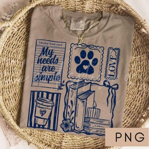 May include: A tan t-shirt with a blue graphic design featuring a library book card with the text "My needs are simple", a paw print with a heart, books, a coffee cup, and the word "LOVE".