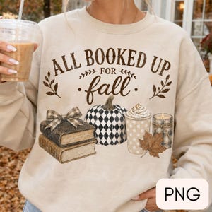 May include: A beige crewneck sweatshirt featuring the brown text ALL BOOKED UP FOR fall, decorated with a stack of two books with a plaid bow, a black and white diamond-patterned pumpkin, a polka-dot coffee cup, a candle, and a brown autumn leaf.