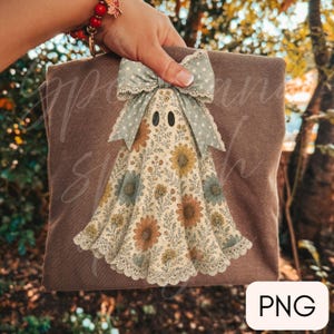 May include: A brown t-shirt featuring a graphic of a ghost adorned with a floral pattern in shades of orange, yellow, and blue. The ghost has a large, light blue polka-dot bow at the top and a white lace trim along the bottom hem.