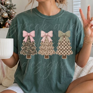May include: A dark green short-sleeve t-shirt featuring a graphic of three stylized Christmas trees. Each tree is topped with a large bow. The first tree has a camouflage pattern, the second a leopard print, and the third a checkered pattern.