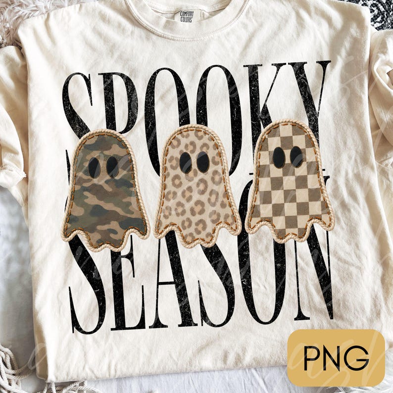 May include: A cream-colored t-shirt featuring the black distressed text SPOOKY SEASON. Three ghost-shaped graphics are centered, each with black oval eyes and a stitched border. The ghosts feature camouflage, leopard print, and brown checkerboard patterns.