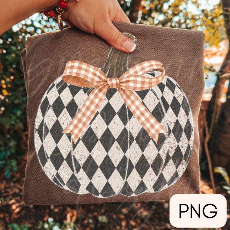 May include: A brown t-shirt featuring a large graphic of a pumpkin with a black and white diamond harlequin pattern. The pumpkin has a dark green stem and is decorated with a tan and white gingham check ribbon bow tied at the top center.