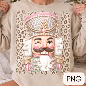 Op de afbeelding: Een beige sweater met ronde hals en een afbeelding van een knipogende notenkraker in een uniform met roze en groen luipaardpatroon, een hoge hoed met gouden accenten en een witte baard, tegen een verticaal paneel met bruin luipaardpatroon.