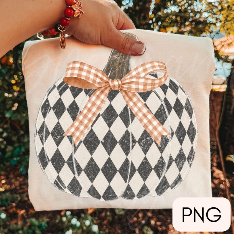 May include: A cream-colored fabric item featuring a large pumpkin graphic with a black and white diamond harlequin pattern. The pumpkin is topped with a light brown and white gingham check ribbon bow tied in a knot at the center of the stem.