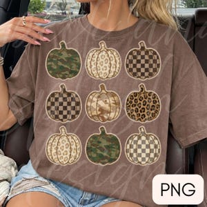 Op de afbeelding: Een bruin T-shirt met korte mouwen voorzien van een raster van negen pompoenillustraties. De pompoenen tonen diverse patronen, waaronder groen camouflage, beige luipaardprint en bruin-witte dambordpatronen met gestikte randen.