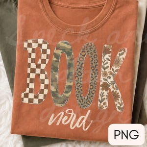 Op de afbeelding: Een roestkleurig t-shirt met het woord BOOK in grote blokletters, elk met een uniek patroon: bruin-wit schaakbord, groen camouflage, bruine luipaardprint en een beige bloemmotief, met het woord nerd in witte schrijfletters.