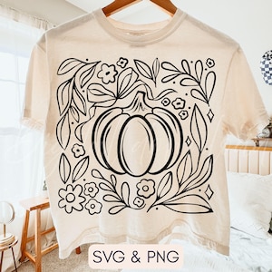 May include: Cream-colored t-shirt featuring a black line art design of a pumpkin surrounded by leaves, flowers, and geometric shapes. The text "SVG & PNG" is at the bottom. The shirt is hanging on a wooden hanger.