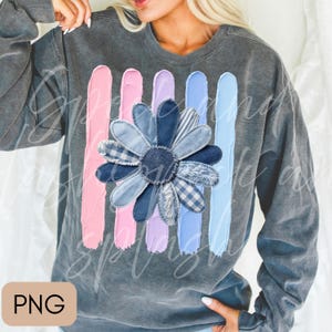 May include: A charcoal grey crewneck sweatshirt featuring a central graphic of a daisy flower composed of various blue denim fabric textures. Behind the flower are five vertical brushstroke stripes in shades of pink, lavender, and light blue.