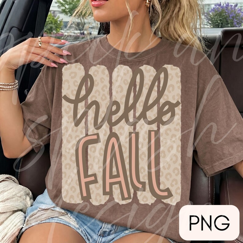 May include: A brown short-sleeve t-shirt featuring a central graphic with five vertical beige stripes overlaid with a leopard print pattern. The text hello FALL is printed in a dark brown and light pink script font across the center of the design.