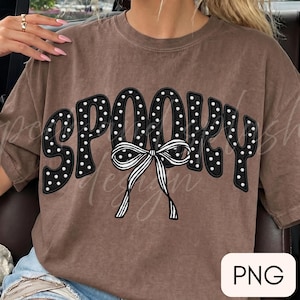 May include: A brown short-sleeved t-shirt featuring the word SPOOKY in black letters with white polka dots. A black and white striped bow is positioned over the center of the word. The text PNG is visible in a white box in the bottom right corner.