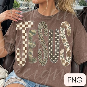 May include: A brown short-sleeve t-shirt featuring the word JESUS in large letters. Each letter has a distinct pattern: a brown and white checkerboard, green camouflage, brown leopard print, white polka dots on brown, and a brown floral print.