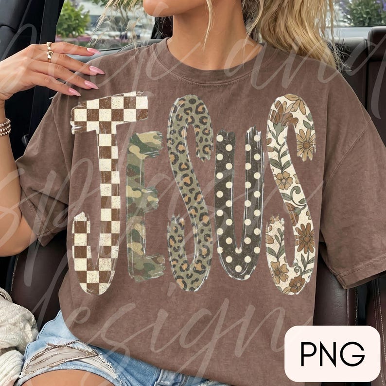 May include: A brown short-sleeve t-shirt featuring the word JESUS in large letters. Each letter has a distinct pattern: a brown and white checkerboard, green camouflage, brown leopard print, white polka dots on brown, and a brown floral print.