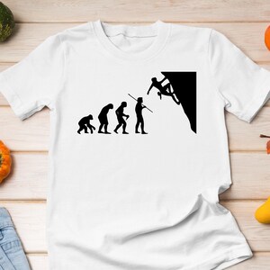 May include: White t-shirt featuring a black silhouette of the evolution of man, culminating in a person rock climbing. The text "Evolution of a Climber" is printed on the shirt.