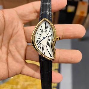 Può includere: Un orologio da polso con cassa asimmetrica color oro e quadrante bianco con numeri romani neri. L'orologio presenta sottili lancette blu ed è fissato a un cinturino in pelle nera con finitura testurizzata. La parola Paris è stampata sul quadrante.
