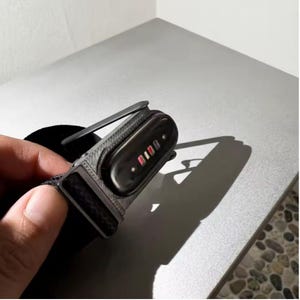 Könnte beinhalten: Ein schwarzer Fitness-Tracker mit strukturiertem Armband und einem glänzenden, ovalen Sensormodul. Die Rückseite des Geräts zeigt vier kleine, rechteckige Ladekontakte in einer Reihe sowie einen seitlich angebrachten Metallclip.