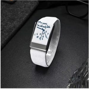 May include: A white fabric wristband featuring a rectangular metal accent and a printed clock face design with the text Who cares I'm already late. The numbers on the clock face are scattered in a non-traditional arrangement with a single hand.