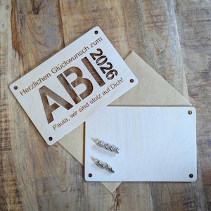 May include: A light brown wooden graduation card with the word "ABI" cut out, the year "2026", and the text "Herzlichen Gl&uuml;ckwunsch zum" and "Paula, wir sind stolz auf Dich!" The card is accompanied by a blank card and a brown envelope.