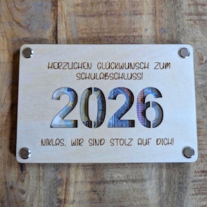 K&ouml;nnte beinhalten: Helle Holzplakette mit Gravur "HERZLICHEN GL&Uuml;CKWUNSCH ZUM SCHULABSCHLUSS!" und "NIKLAS, WIR SIND STOLZ AUF DICH!". Das Jahr "2026" ist ausgeschnitten und zeigt Geld. Vier silberfarbene Nieten befestigen die Plakette.