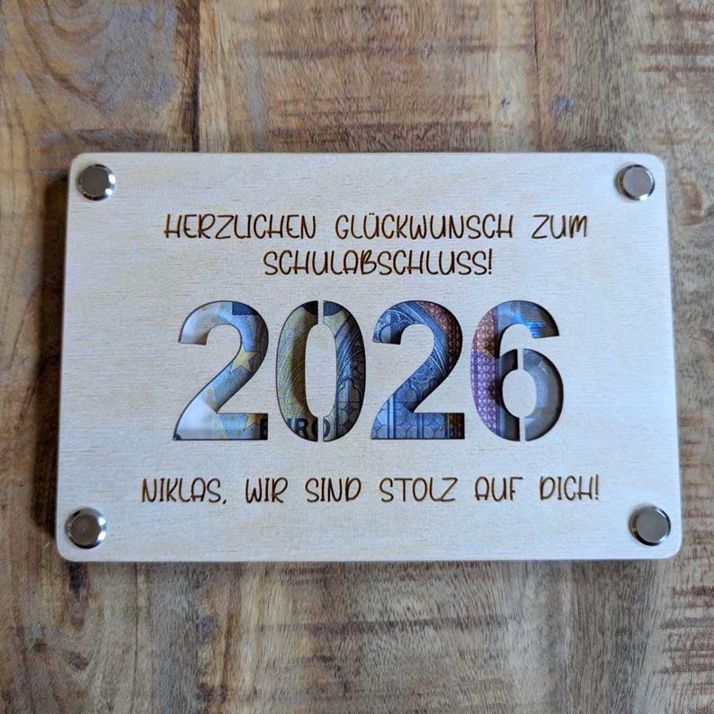 K&ouml;nnte beinhalten: Helle Holzplakette mit Gravur "HERZLICHEN GL&Uuml;CKWUNSCH ZUM SCHULABSCHLUSS!" und "NIKLAS, WIR SIND STOLZ AUF DICH!". Das Jahr "2026" ist ausgeschnitten und zeigt Geld. Vier silberfarbene Nieten befestigen die Plakette.