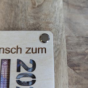May include: A light-coloured wooden plaque with the engraved number "2022" and German text. A silver-coloured metal button is affixed to the top corner. The plaque is partially visible, resting on a wooden surface.