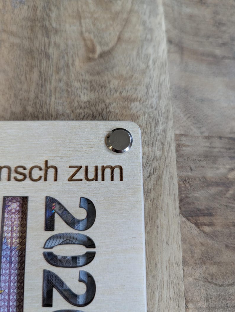 May include: A light-coloured wooden plaque with the engraved number "2022" and German text. A silver-coloured metal button is affixed to the top corner. The plaque is partially visible, resting on a wooden surface.