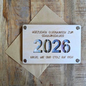 K&ouml;nnte beinhalten: Eine Holz-Abschlusskarte mit dem ausgeschnittenen Jahr 2026, das farbenfrohe W&auml;hrungen zeigt. Die Karte tr&auml;gt die Aufschrift "HERZLICHEN GL&Uuml;CKWUNSCH ZUM SCHULABSCHLUSS!" und "NIKLAS, WIR SIND STOLZ AUF DICH!" und ist an einem braunen Umschlag befestigt.