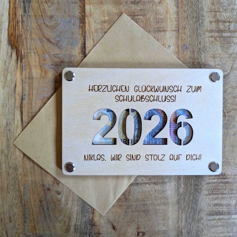 K&ouml;nnte beinhalten: Eine Holz-Abschlusskarte mit dem ausgeschnittenen Jahr 2026, das farbenfrohe W&auml;hrungen zeigt. Die Karte tr&auml;gt die Aufschrift "HERZLICHEN GL&Uuml;CKWUNSCH ZUM SCHULABSCHLUSS!" und "NIKLAS, WIR SIND STOLZ AUF DICH!" und ist an einem braunen Umschlag befestigt.