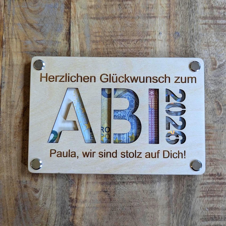 May include: A light-coloured wooden plaque with the word "ABI" cut out, revealing currency. The year "2026" is also cut out. The text "Herzlichen Gl&uuml;ckwunsch zum" and "Paula, wir sind stolz auf Dich!" are engraved. Four silver rivets secure the plaque.