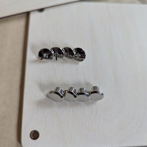 K&ouml;nnte beinhalten: Nahaufnahme von gl&auml;nzenden silbernen Metallschrauben und -nieten auf einer hellen Holzoberfl&auml;che. Die Schrauben haben eine kreuzf&ouml;rmige Vertiefung, w&auml;hrend die Nieten einen flachen, abgerundeten Kopf haben. Das Holz hat eine nat&uuml;rliche Maserung.
