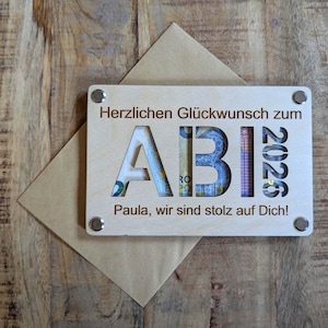 May include: A wooden graduation gift with the word "ABI" cut out, revealing money inside. The year "2020" is also cut out. The text "Herzlichen Gl&uuml;ckwunsch zum" and "Paula, wir sind stolz auf Dich!" are engraved on the wood. The gift is on a wooden surface.