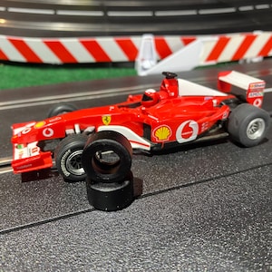 May include: A red and white toy race car with a Shell logo and the number 6 on the side. The car is on a black track with a red and white barrier in the background. There are three black tires next to the car.