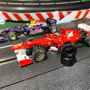May include: Two toy Formula 1 race cars on a track. The red car has the number 5 and the Ferrari logo. The other car is blue with the Red Bull logo. The track has white and red barriers.
