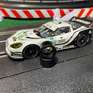 May include: A white and black race car model with sponsor decals and the number 72. The model has a rear spoiler and is on a track with two black tires in front. The car has the Dunlop logo.