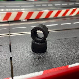 May include: Two black rubber tyres stacked on a grey surface, possibly for a slot car. The tyres have a circular design with a hole in the centre. The background includes a race track with red and white barriers.