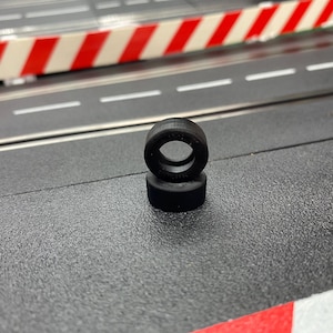 May include: Two small, black rubber tires stacked on a textured gray surface. The tires have a circular opening in the center. The background includes a race track with white lines and a red and white barrier.