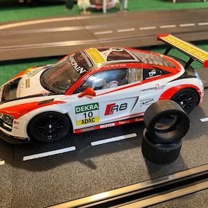 Puede incluir: Un coche de carreras de juguete blanco, rojo y negro con el número 10 y el texto "ADAC" en el lateral. El coche está en una pista con dos neumáticos de repuesto cerca. El coche tiene el texto "R8" en el lateral.