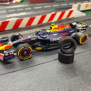 May include: A navy blue, yellow, and red Formula 1 model race car with sponsor branding, and two spare black tyres. The car sits on a grey track, with red and white barriers. The model includes intricate details.