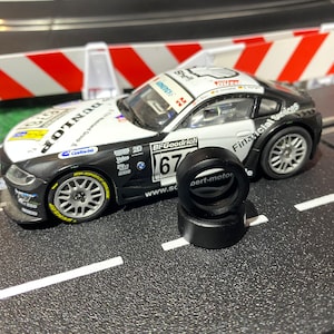 Puede incluir: Un coche de carreras de juguete negro y blanco con las palabras "Financial Services" y "BF Goodrich" en el lateral. El coche está en una pista negra con líneas blancas. Hay tres neumáticos negros en primer plano.