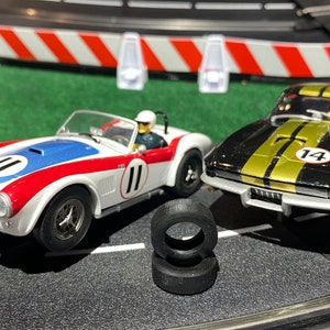 May include: Two toy race cars on a track. The car on the left is white with red and blue accents and the number 11. The car on the right is black with a gold stripe and the number 14. There are two black tires in the foreground.